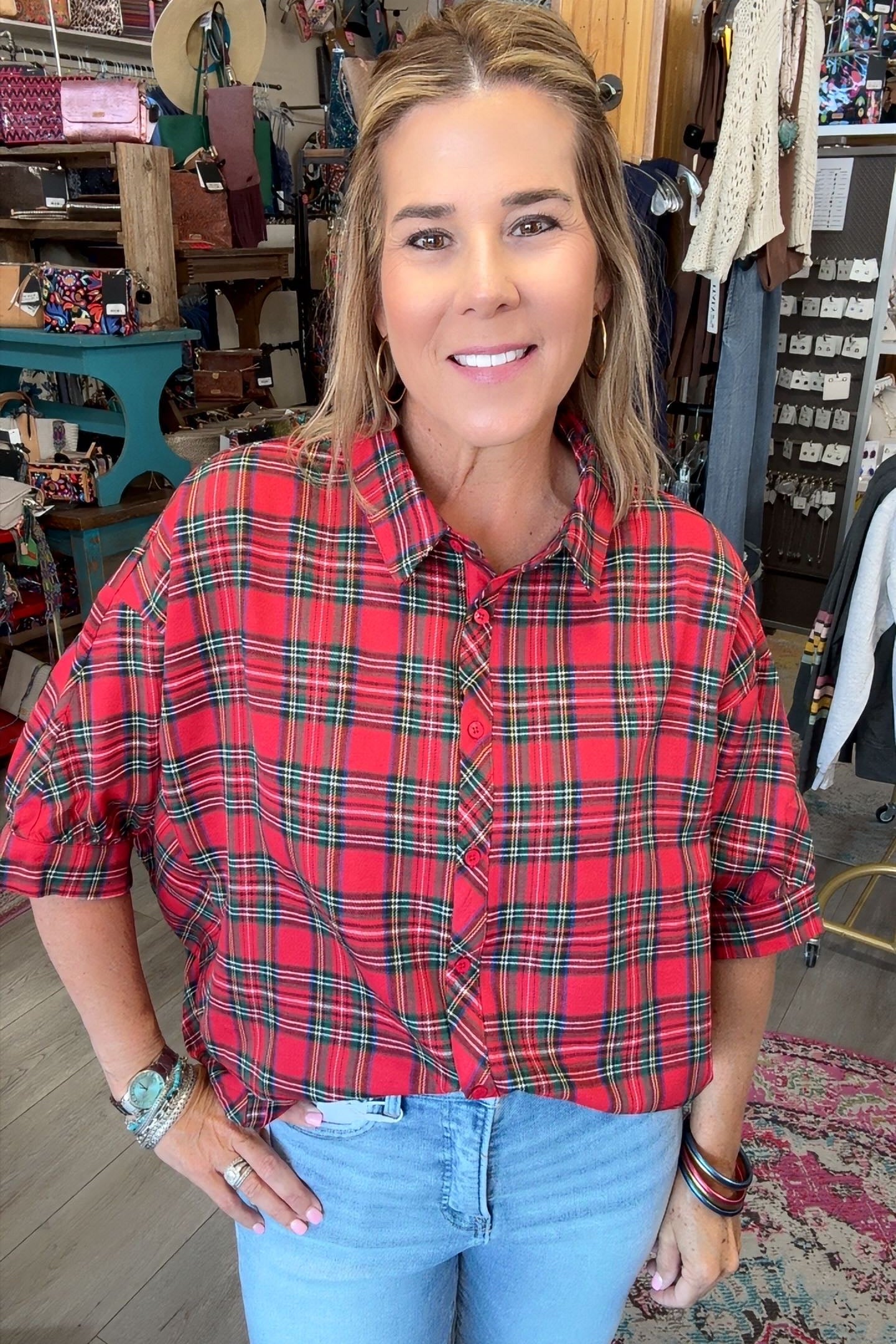 A woman wearing a red and green plaid short-sleeve button-up shirt with light blue jeans, standing in a retail store.