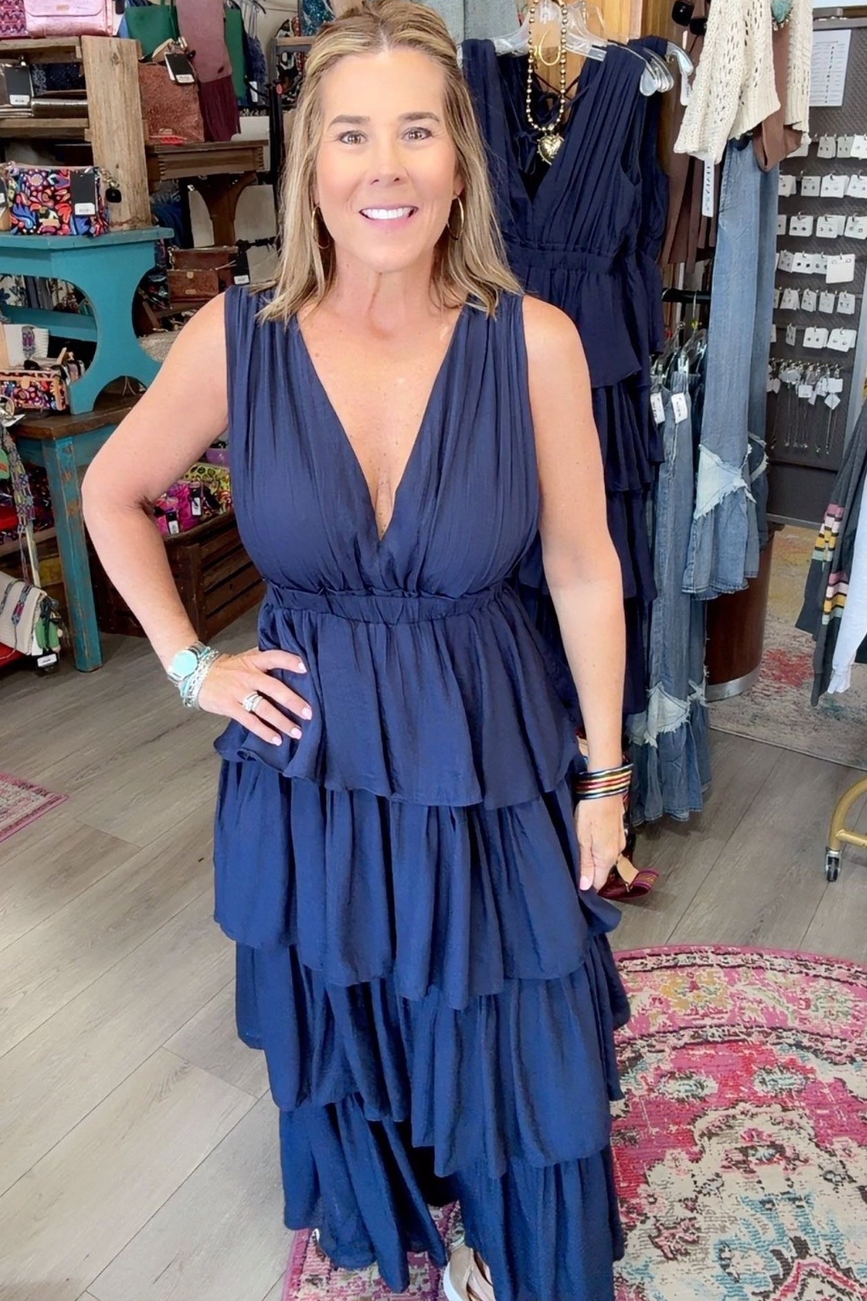 A woman modeling a sleeveless navy blue tiered maxi dress in a boutique setting with clothing racks and shelves in the background.
