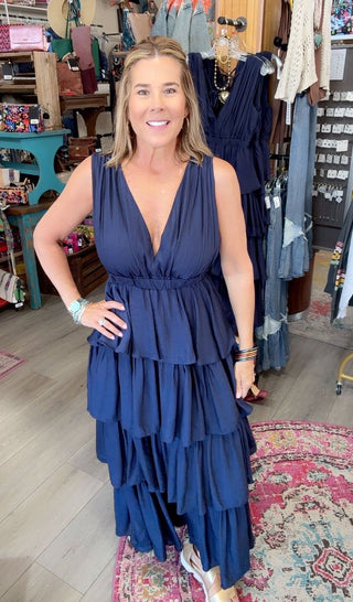 A woman modeling a sleeveless navy blue tiered maxi dress in a boutique setting with clothing racks and shelves in the background.