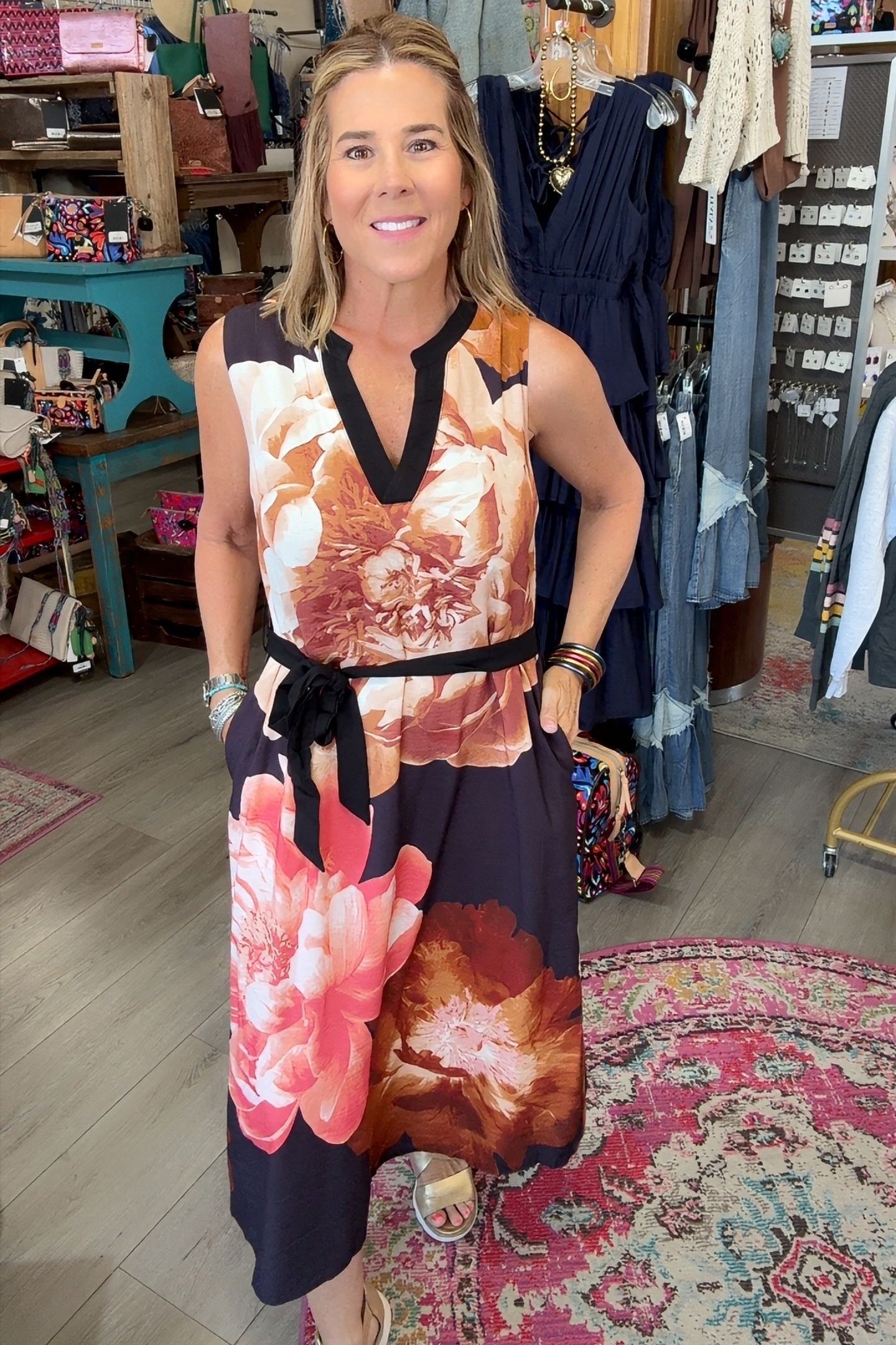 A woman modeling a sleeveless V-neck dress with a large floral print in pink, orange, and dark navy, standing inside a boutique shop.