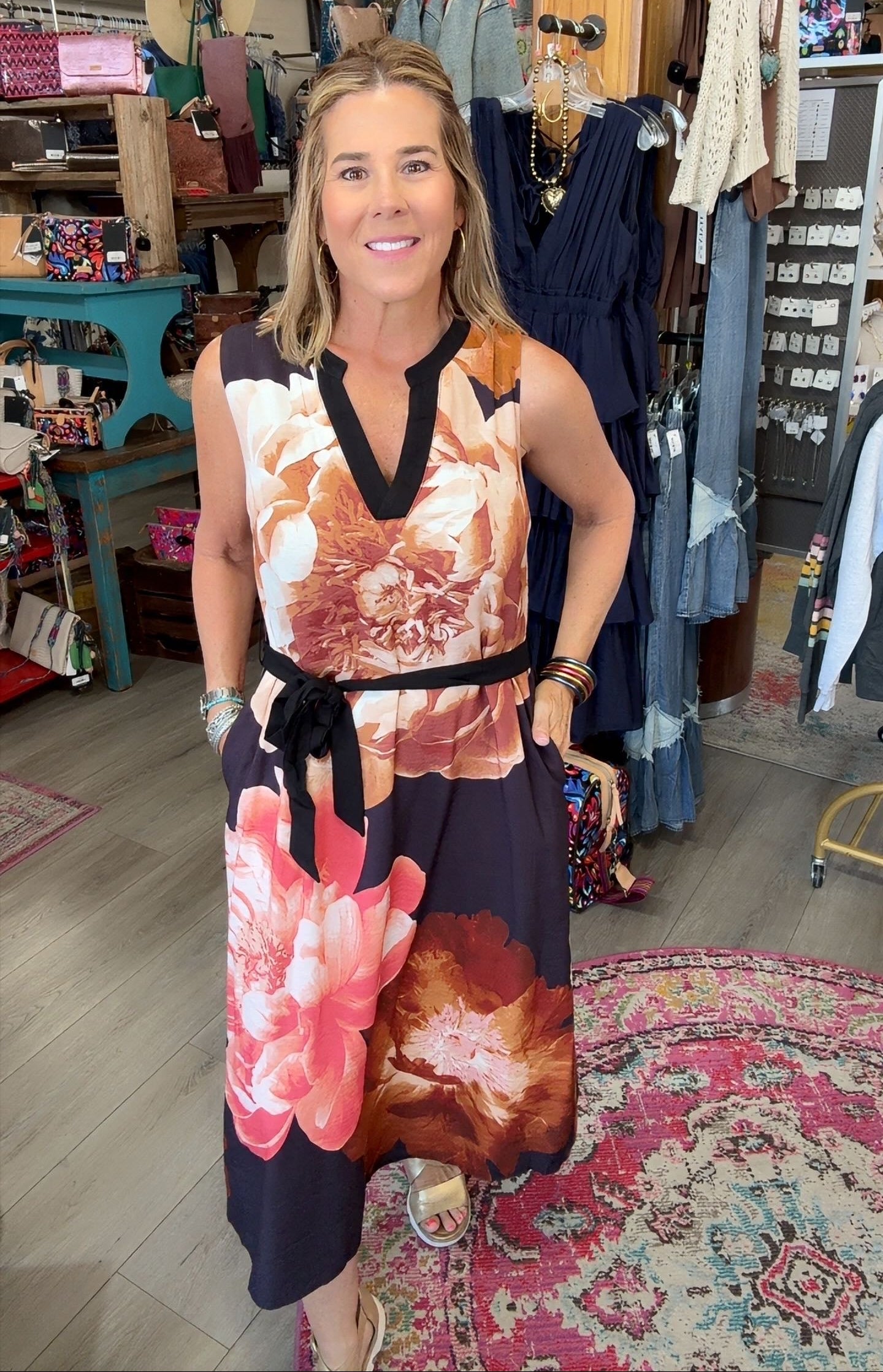 A woman modeling a sleeveless V-neck dress with a large floral print in pink, orange, and dark navy, standing inside a boutique shop.