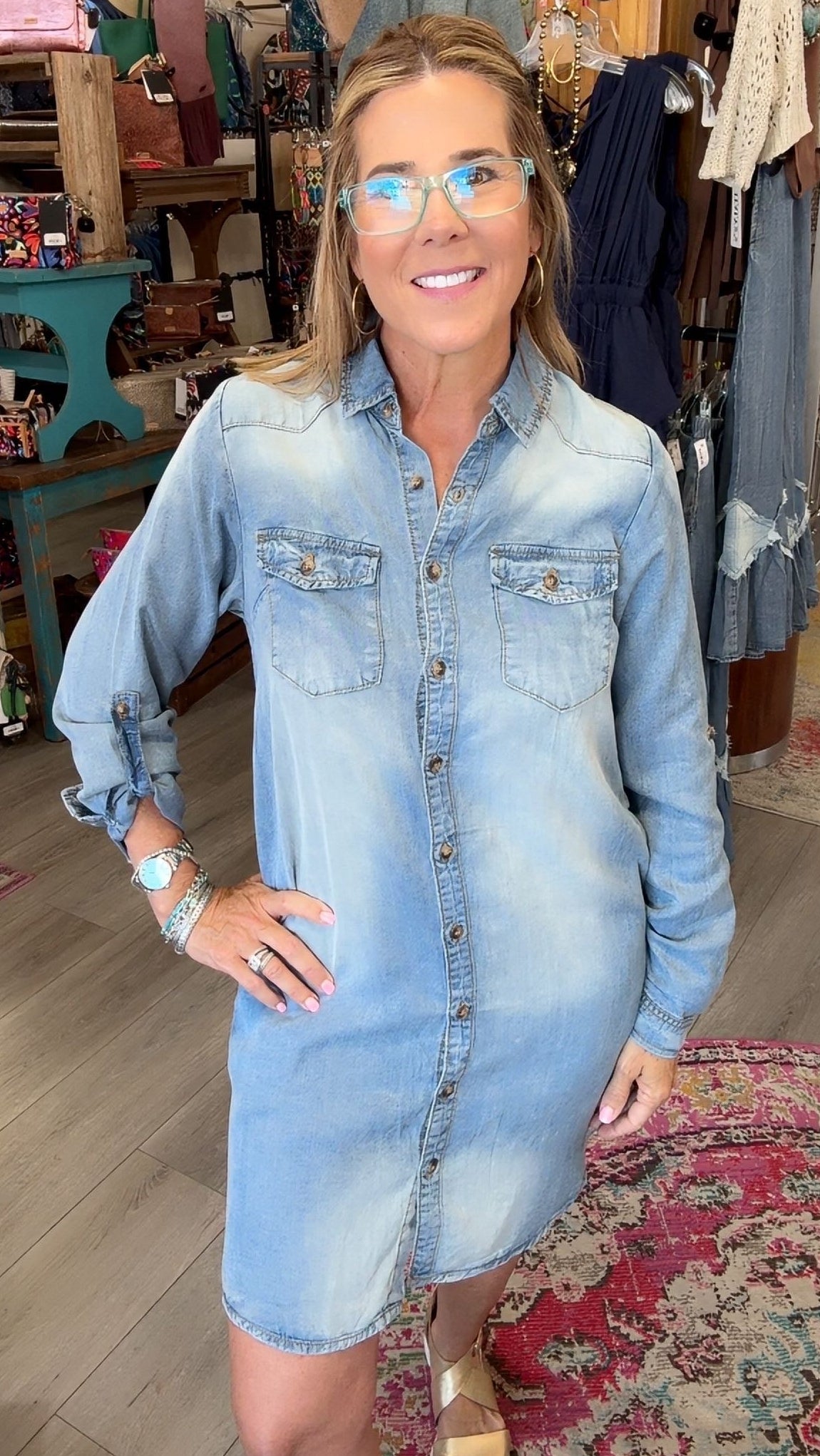 A woman wearing a light blue denim shirt dress stands inside a retail boutique. The dress has a faded wash, a button-down front, and two chest pockets.