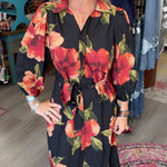 A woman modeling a black long-sleeve midi dress with a vibrant red and orange floral print, standing inside a boutique shop.