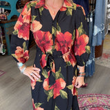 A woman modeling a black long-sleeve midi dress with a vibrant red and orange floral print, standing inside a boutique shop.