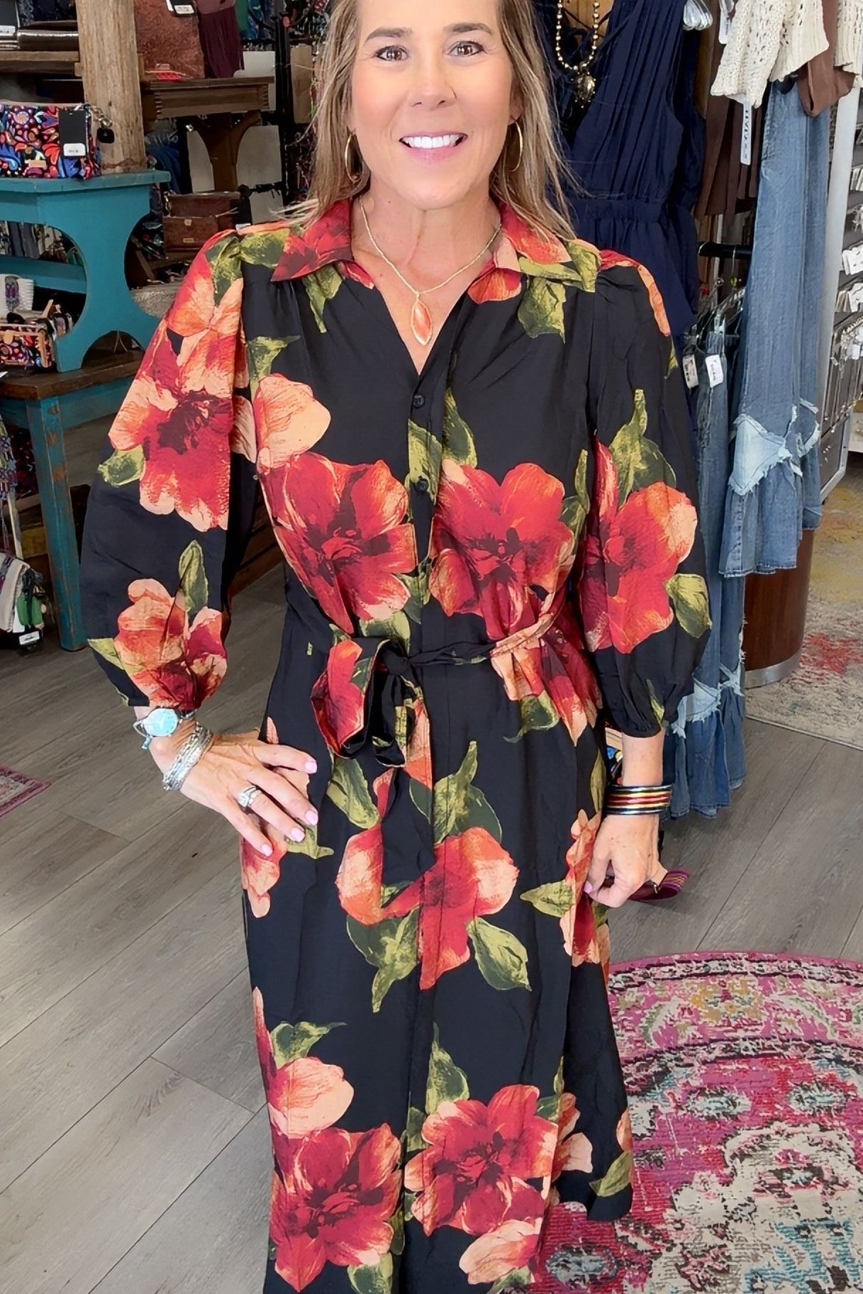 A woman modeling a black long-sleeve midi dress with a vibrant red and orange floral print, standing inside a boutique shop.