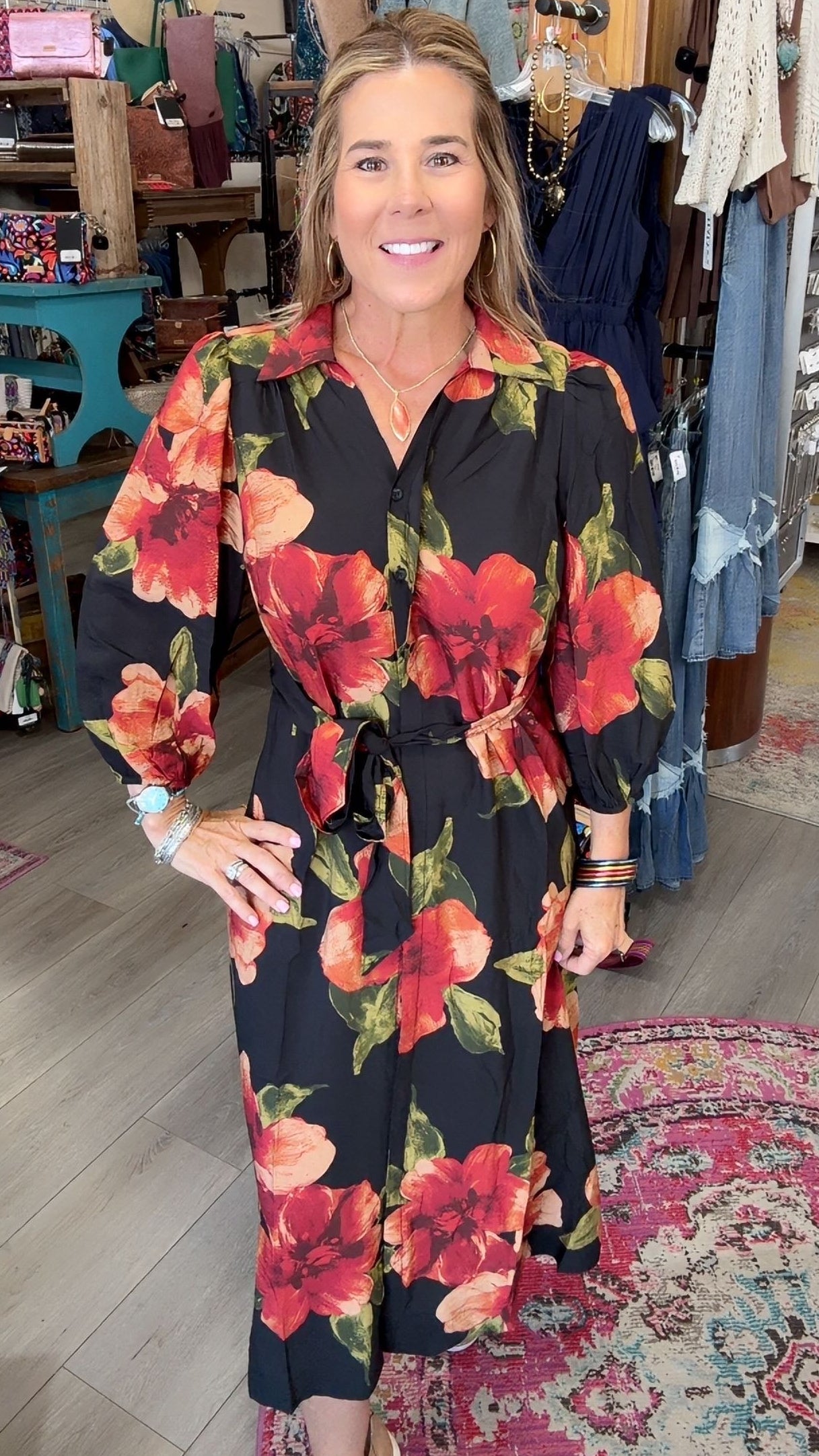 A woman modeling a black long-sleeve midi dress with a vibrant red and orange floral print, standing inside a boutique shop.