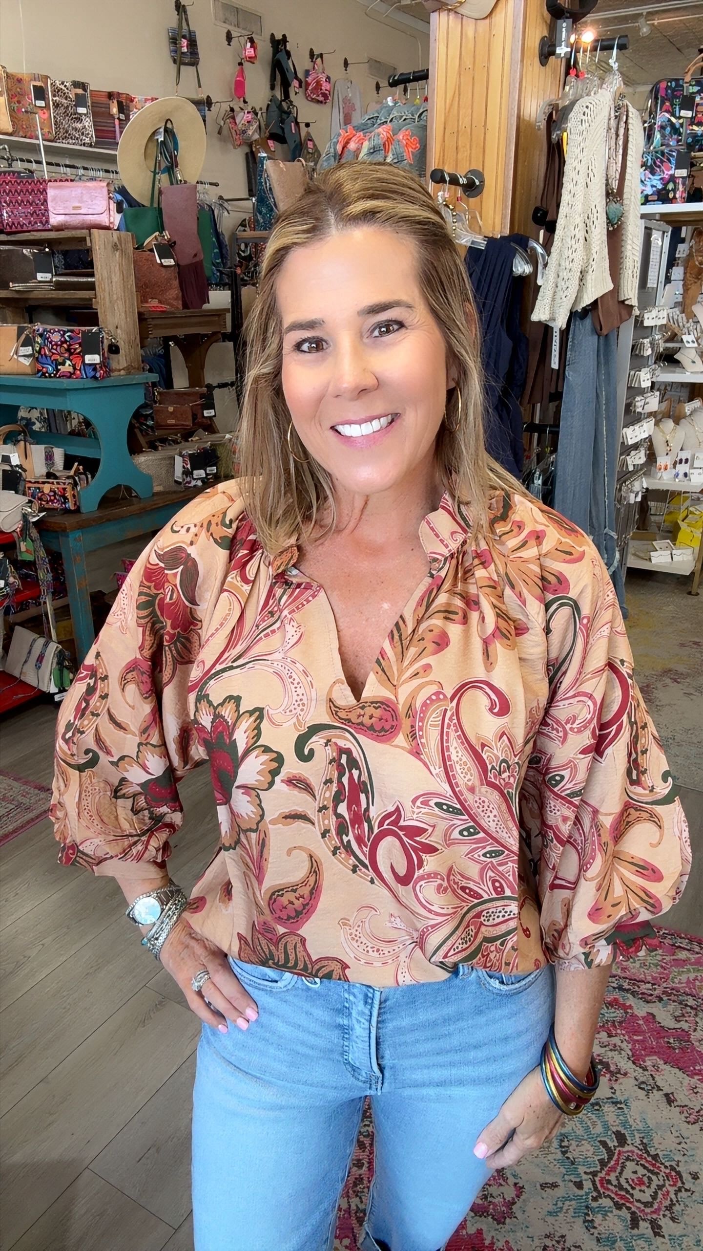 A smiling woman with blonde hair stands in a retail store wearing a beige button-up blouse with a red and green paisley pattern, paired with light blue jeans.