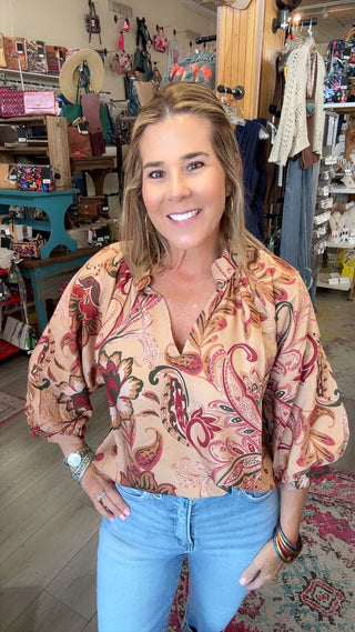 A smiling woman with blonde hair stands in a retail store wearing a beige button-up blouse with a red and green paisley pattern, paired with light blue jeans.