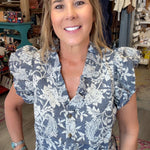 A smiling woman with blonde hair stands in a retail store wearing a slate blue and white floral print blouse with puff sleeves and light blue jeans.