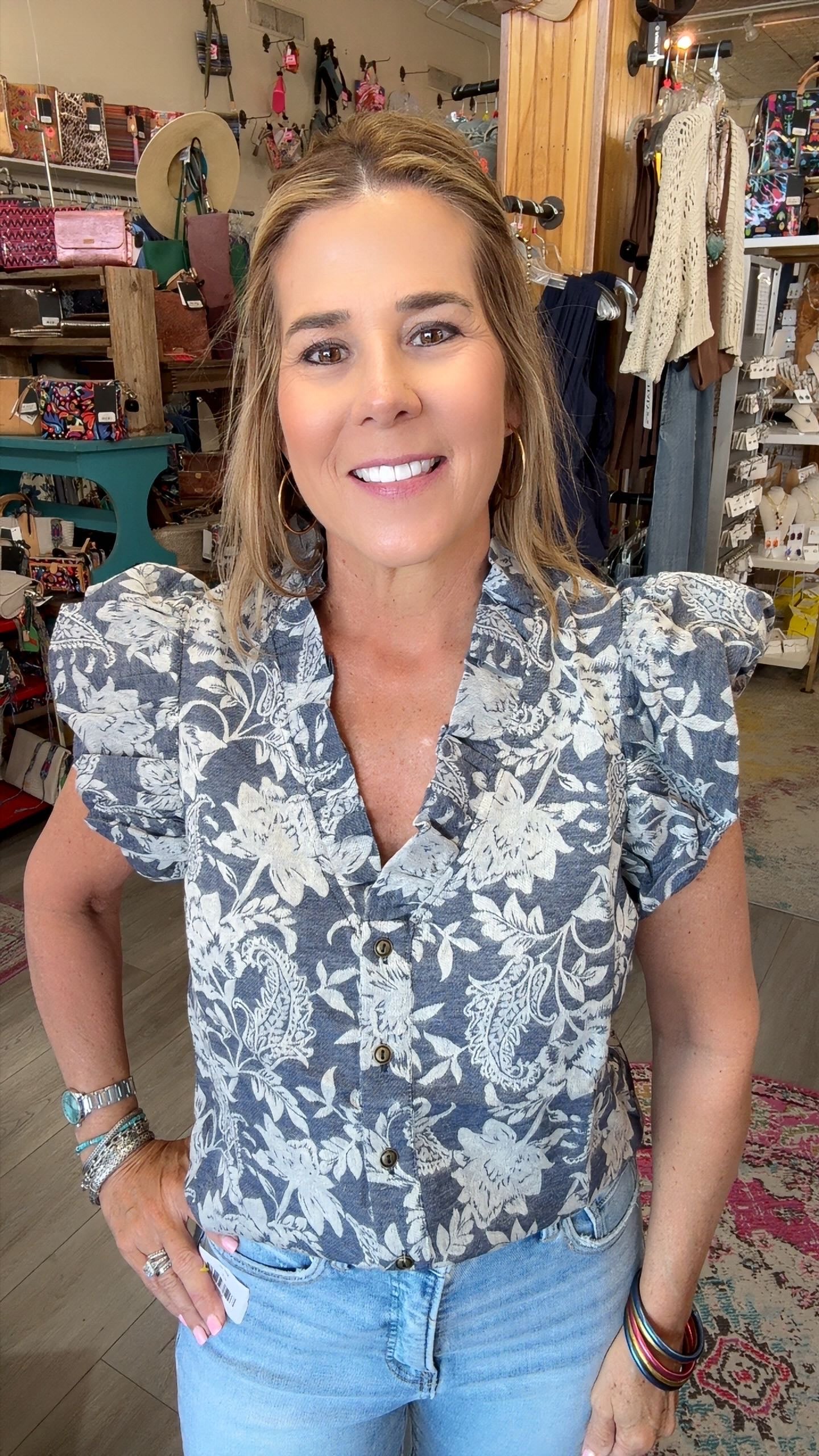 A smiling woman with blonde hair stands in a retail store wearing a slate blue and white floral print blouse with puff sleeves and light blue jeans.