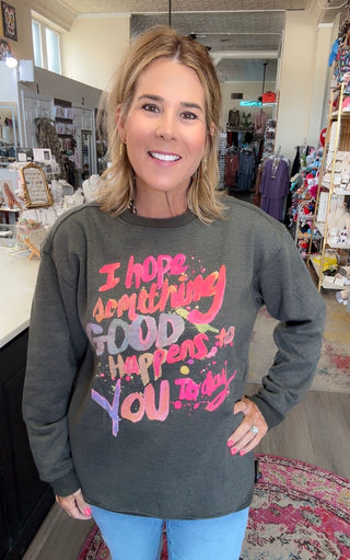 A woman wearing a dark gray graphic sweatshirt with colorful text that reads 'I hope something good happens to you today' in pink, orange, and purple, standing inside a retail store.