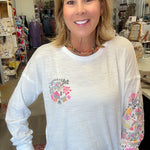 A smiling woman with blonde hair stands in a boutique wearing a white long-sleeve tunic top with a pink floral heart design on the chest and a floral pattern down one sleeve.