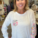 A smiling woman with blonde hair stands in a boutique wearing a white long-sleeve tunic top with a pink floral heart design on the chest and a floral pattern down one sleeve.