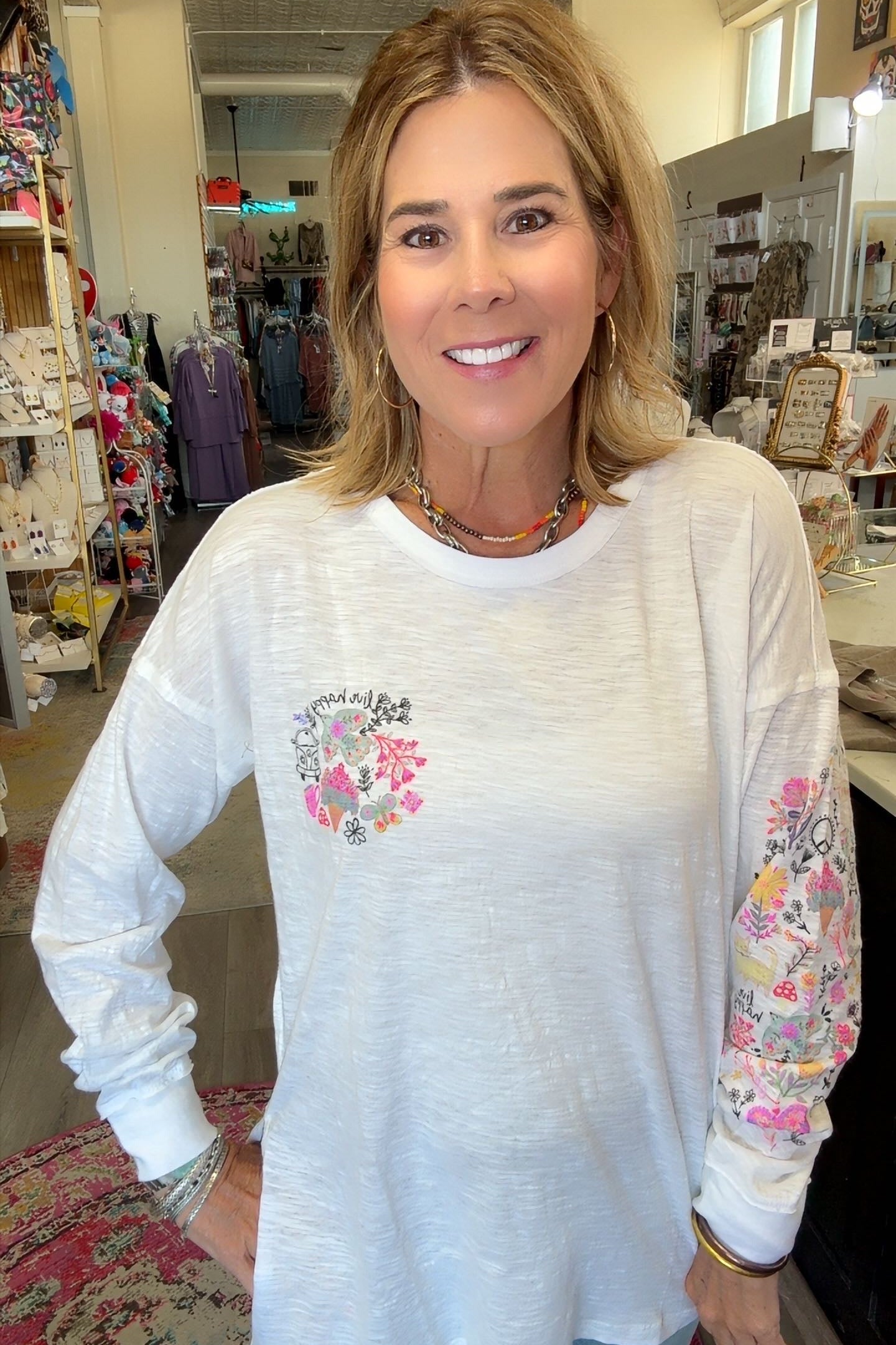 A smiling woman with blonde hair stands in a boutique wearing a white long-sleeve tunic top with a pink floral heart design on the chest and a floral pattern down one sleeve.