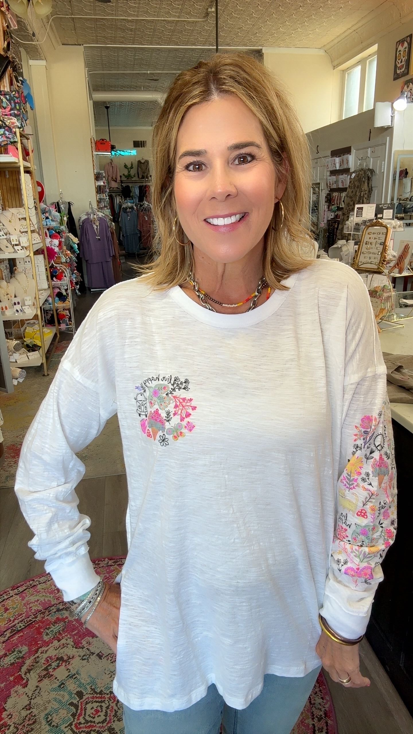 A smiling woman with blonde hair stands in a boutique wearing a white long-sleeve tunic top with a pink floral heart design on the chest and a floral pattern down one sleeve.