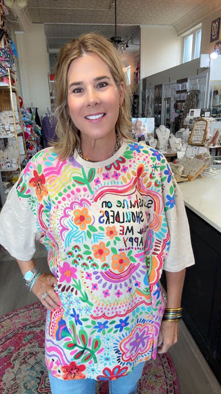 A woman smiling in a colorful, bohemian-style graphic t-shirt with white sleeves, standing inside a retail shop.