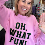 A woman smiling and waving while wearing a pink crewneck sweatshirt with the text 'OH, WHAT FUN!' in black glitter-style lettering, paired with blue jeans in a retail store setting.