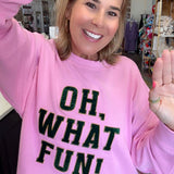 A woman smiling and waving while wearing a pink crewneck sweatshirt with the text 'OH, WHAT FUN!' in black glitter-style lettering, paired with blue jeans in a retail store setting.