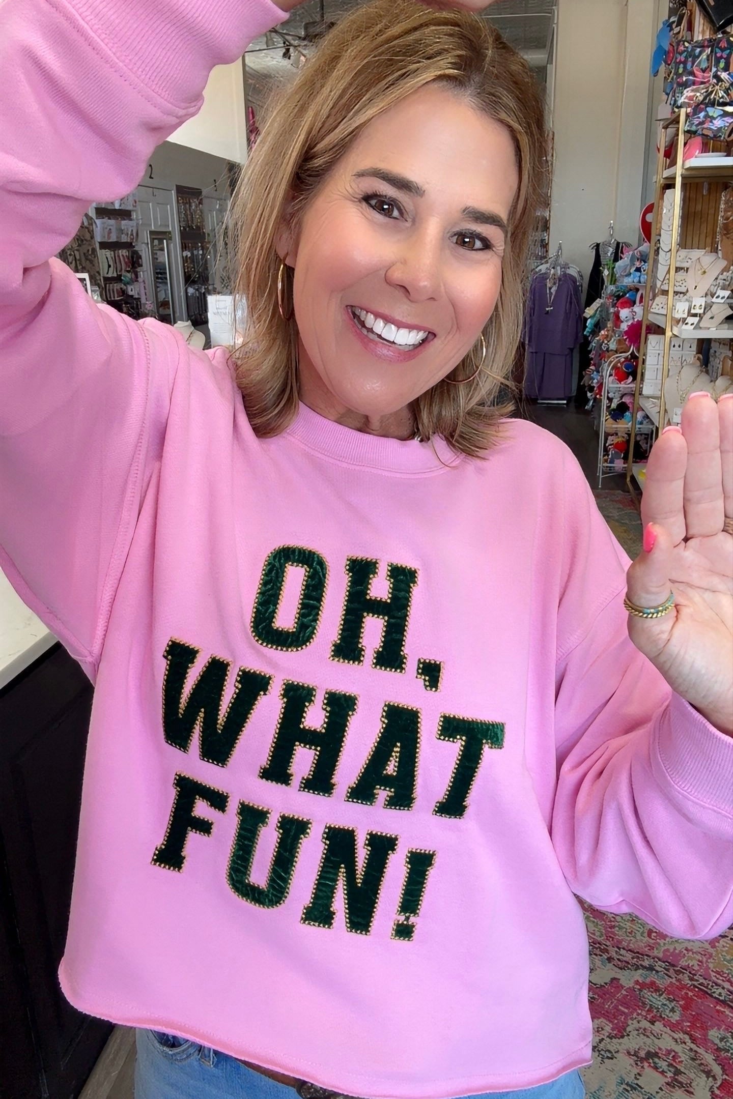 A woman smiling and waving while wearing a pink crewneck sweatshirt with the text 'OH, WHAT FUN!' in black glitter-style lettering, paired with blue jeans in a retail store setting.