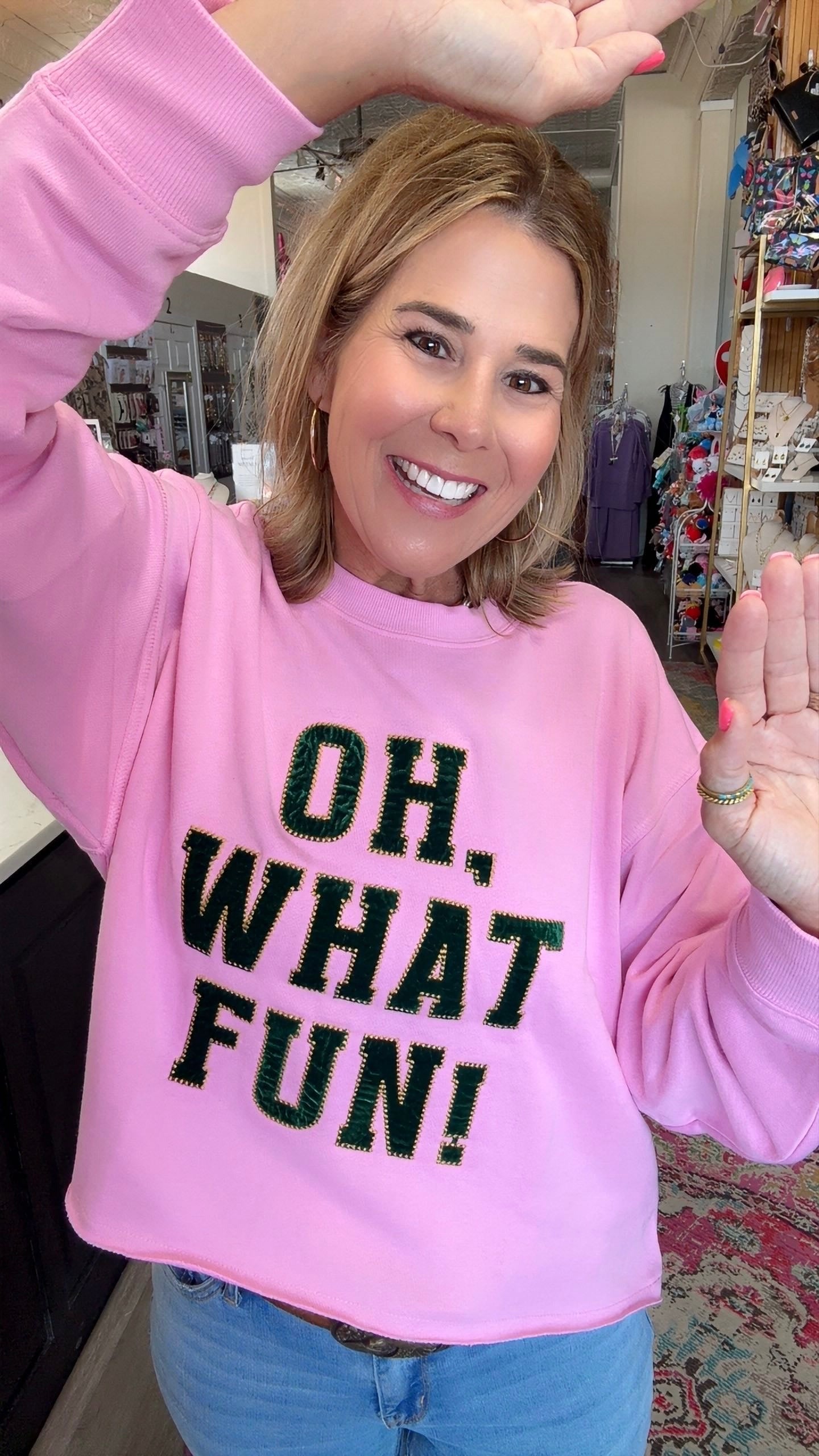 A woman smiling and waving while wearing a pink crewneck sweatshirt with the text 'OH, WHAT FUN!' in black glitter-style lettering, paired with blue jeans in a retail store setting.