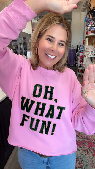 A woman smiling and waving while wearing a pink crewneck sweatshirt with the text 'OH, WHAT FUN!' in black glitter-style lettering, paired with blue jeans in a retail store setting.
