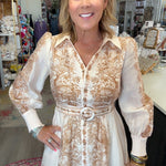 A woman modeling a long-sleeve button-front dress with a beige and white toile pattern, standing inside a retail store.