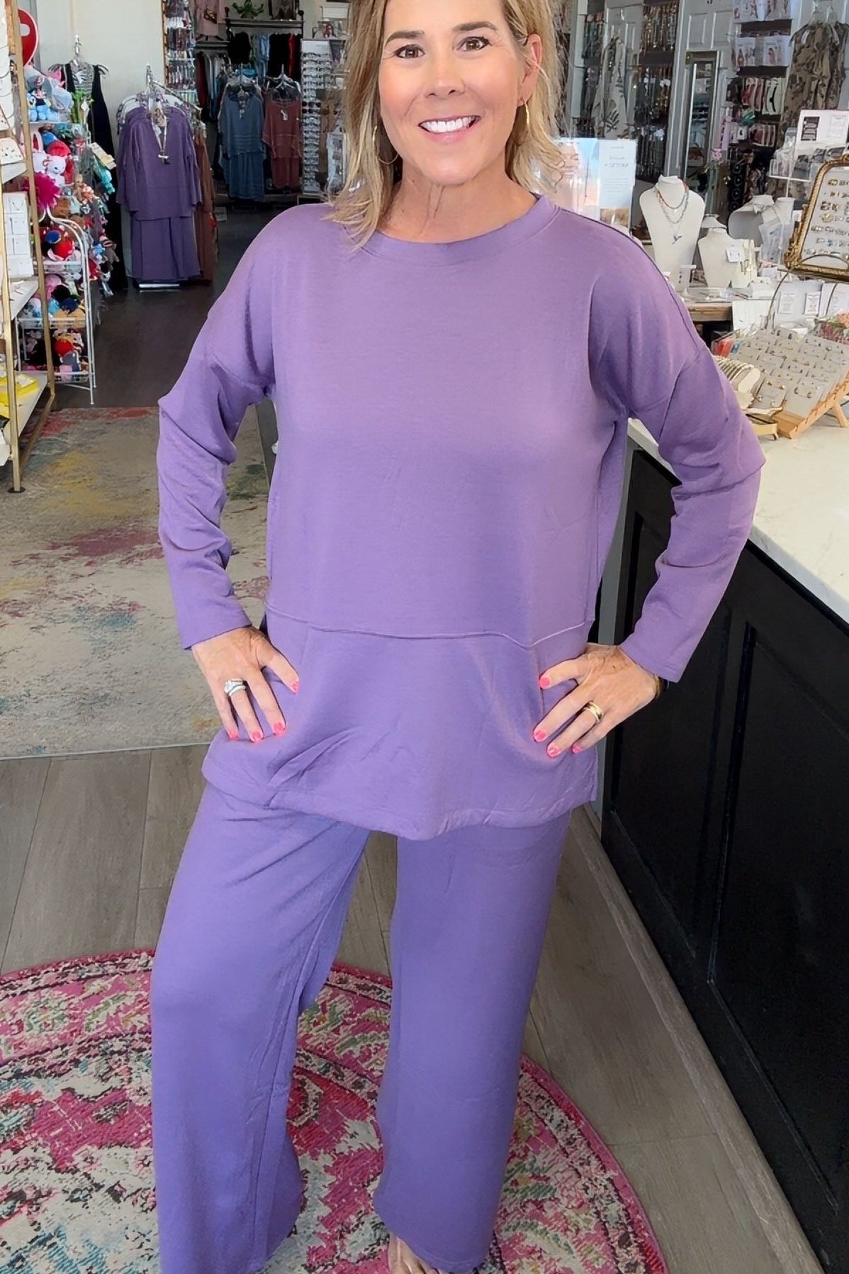 A woman modeling a matching lavender lounge set consisting of a long-sleeve top and wide-leg pants, standing in a retail store.