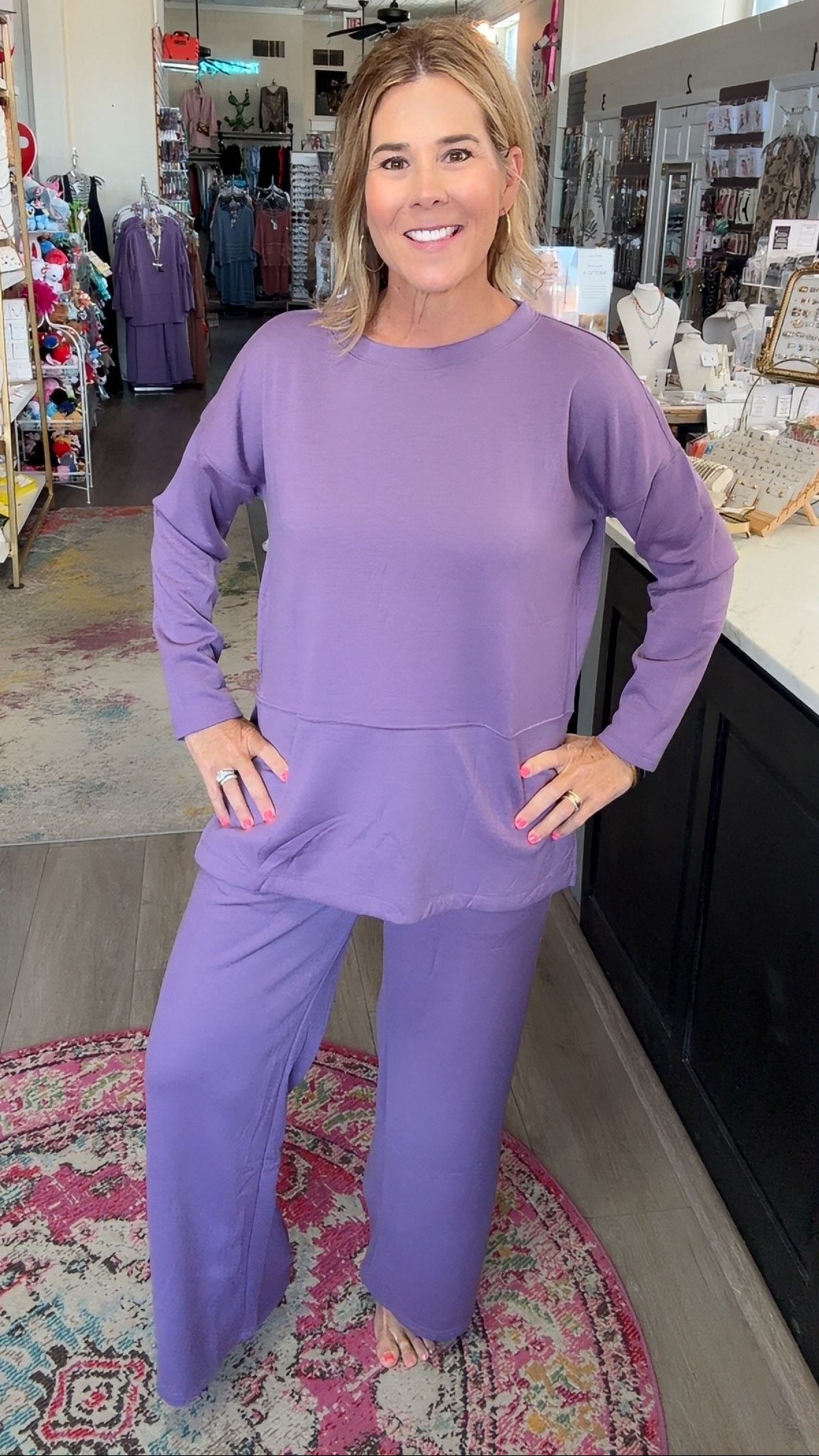A woman modeling a matching lavender lounge set consisting of a long-sleeve top and wide-leg pants, standing in a retail store.