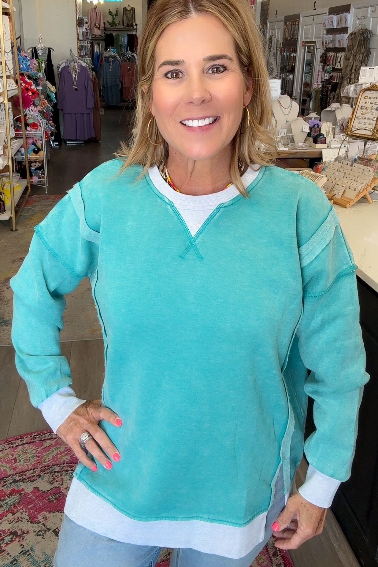 A woman modeling a turquoise long-sleeve v-neck sweater with white trim and a white undershirt, standing inside a retail boutique.