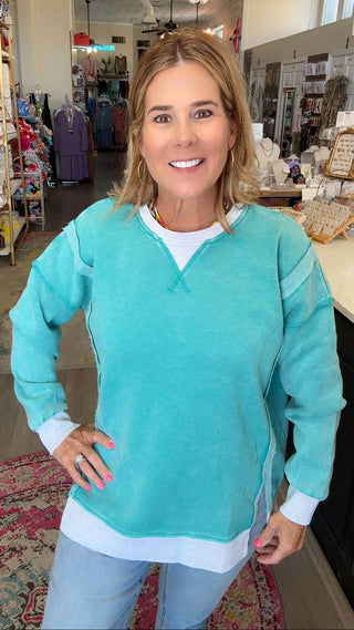 A woman modeling a turquoise long-sleeve v-neck sweater with white trim and a white undershirt, standing inside a retail boutique.