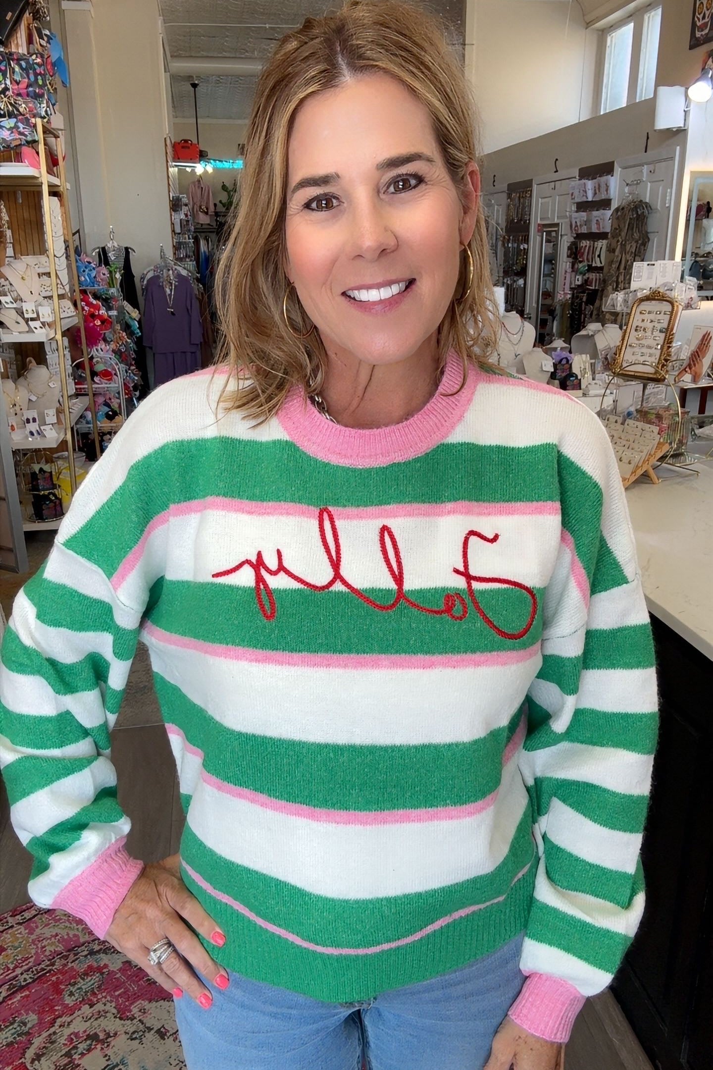 A woman wearing a green, white, and pink striped sweater with 'Folly' embroidered in red across the chest, standing in a retail store.