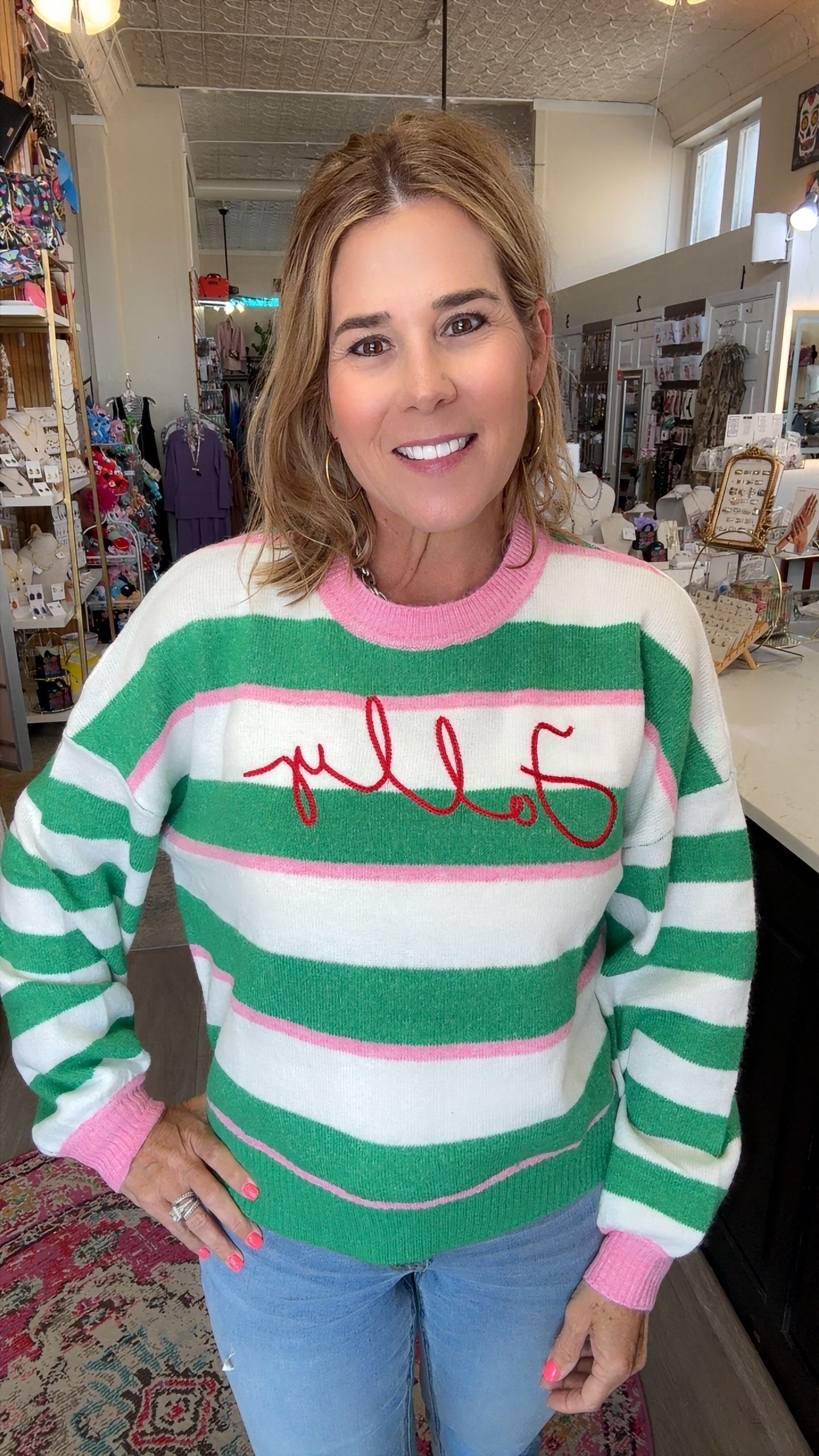 A woman wearing a green, white, and pink striped sweater with 'Folly' embroidered in red across the chest, standing in a retail store.