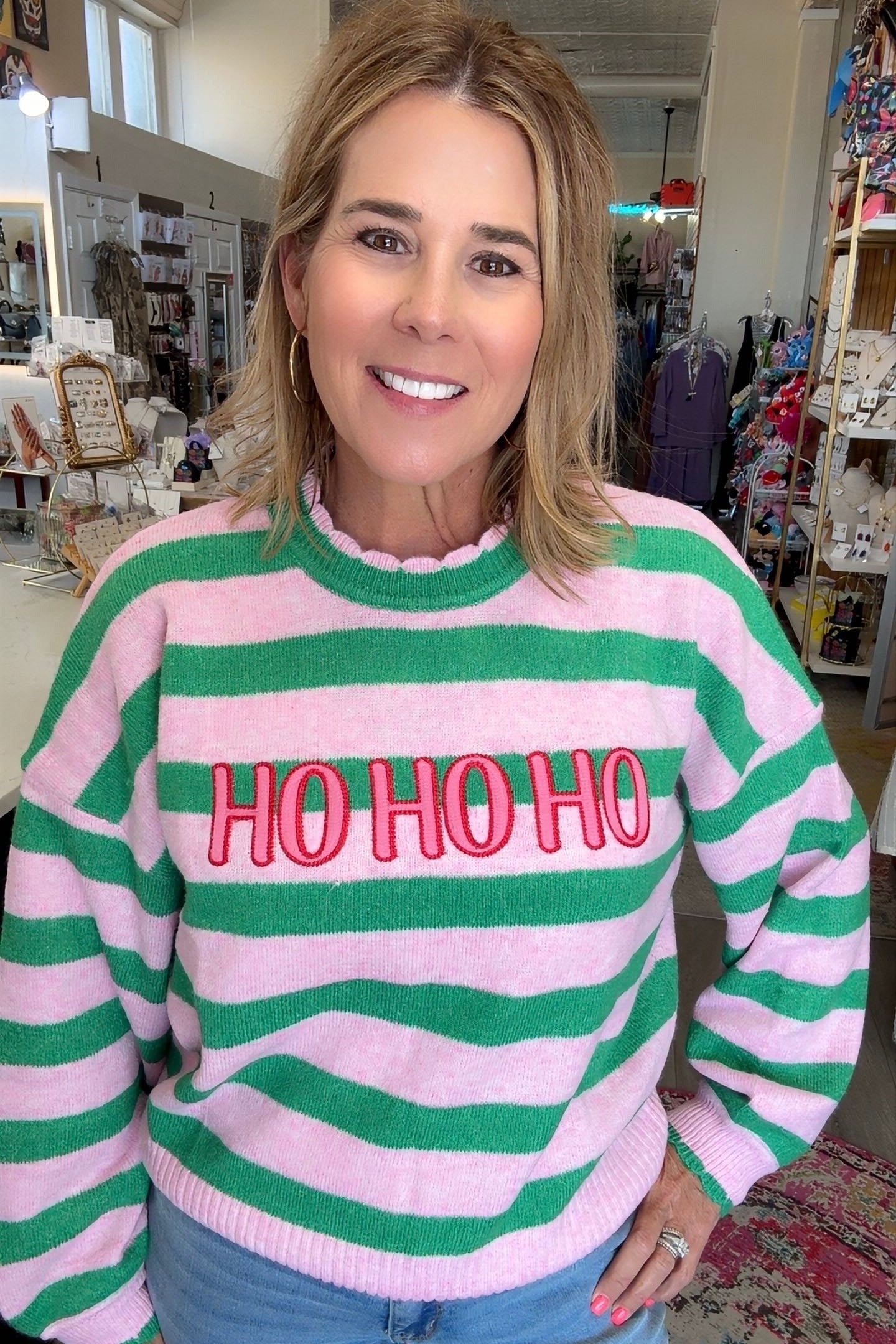 A woman wearing a pink and green horizontally striped sweater with 'HO HO HO' embroidered in red across the chest, paired with light blue jeans, standing inside a retail store.