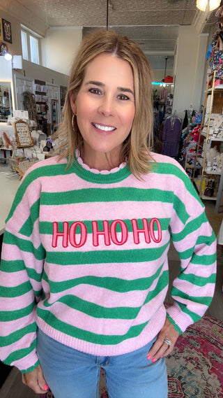 A woman wearing a pink and green horizontally striped sweater with 'HO HO HO' embroidered in red across the chest, paired with light blue jeans, standing inside a retail store.