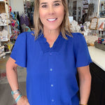 A woman with blonde hair smiling in a boutique, wearing a bright royal blue short-sleeve button-up blouse with a textured collar and black pants.