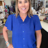 A woman with blonde hair smiling in a boutique, wearing a bright royal blue short-sleeve button-up blouse with a textured collar and black pants.