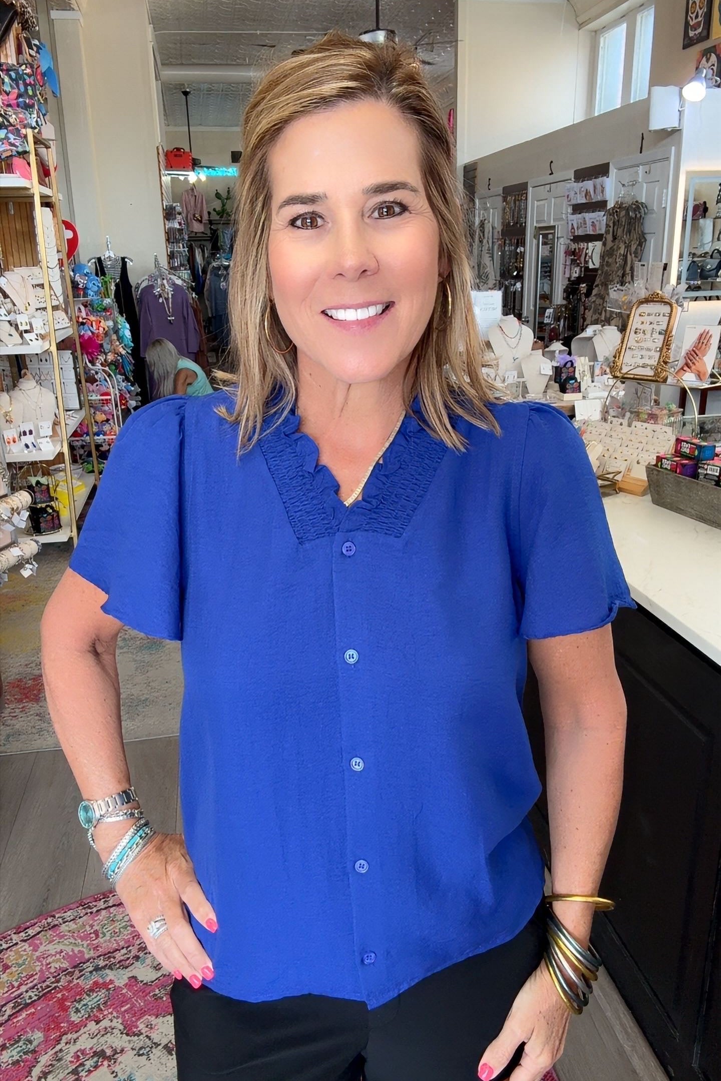 A woman with blonde hair smiling in a boutique, wearing a bright royal blue short-sleeve button-up blouse with a textured collar and black pants.