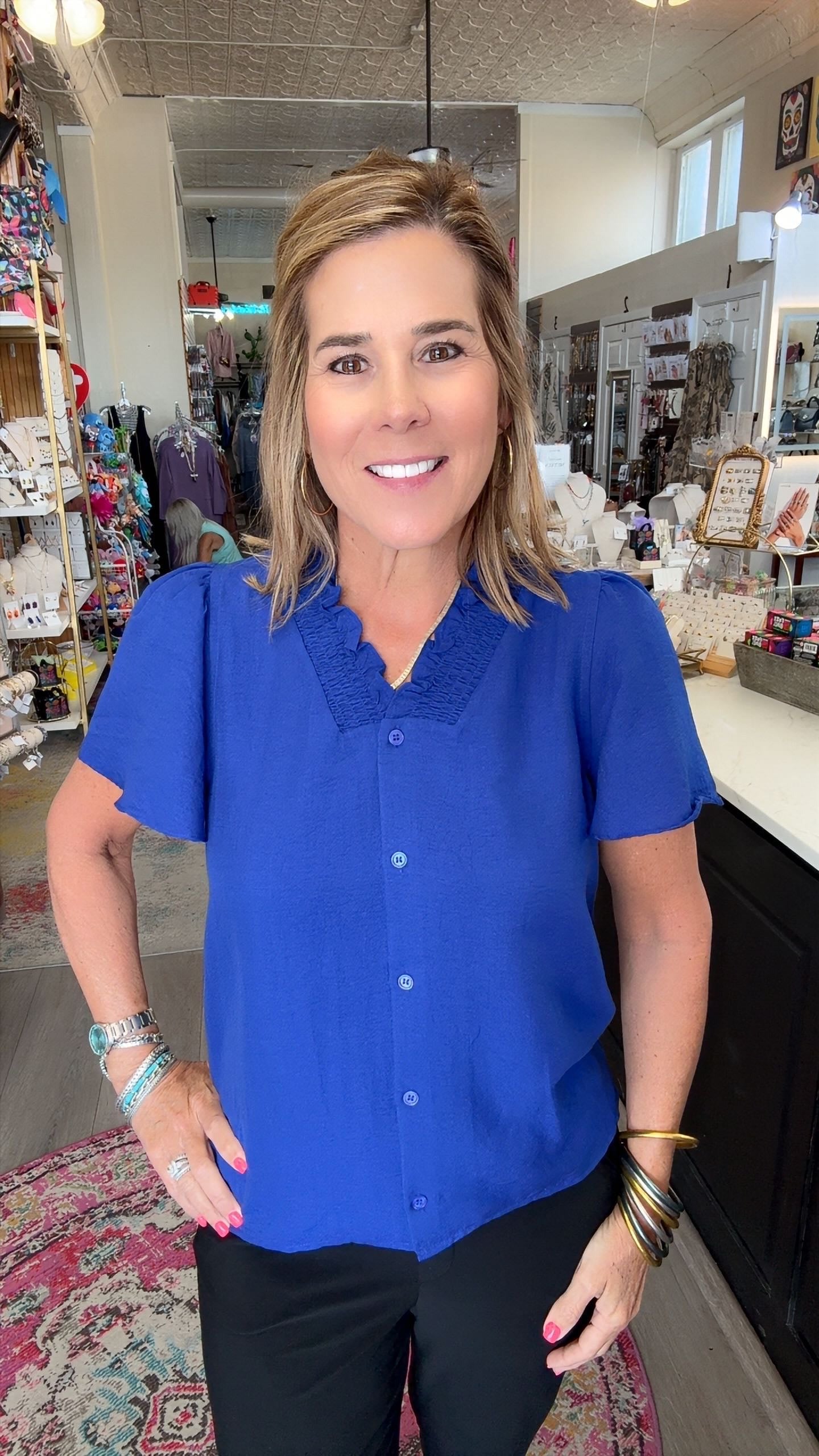 A woman with blonde hair smiling in a boutique, wearing a bright royal blue short-sleeve button-up blouse with a textured collar and black pants.