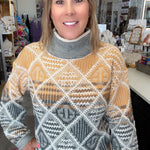A woman wearing a patterned turtleneck sweater in a retail store setting. The sweater features a geometric diamond pattern in mustard yellow, gray, white, and brown.
