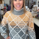 A woman wearing a patterned turtleneck sweater in a retail store setting. The sweater features a geometric diamond pattern in mustard yellow, gray, white, and brown.