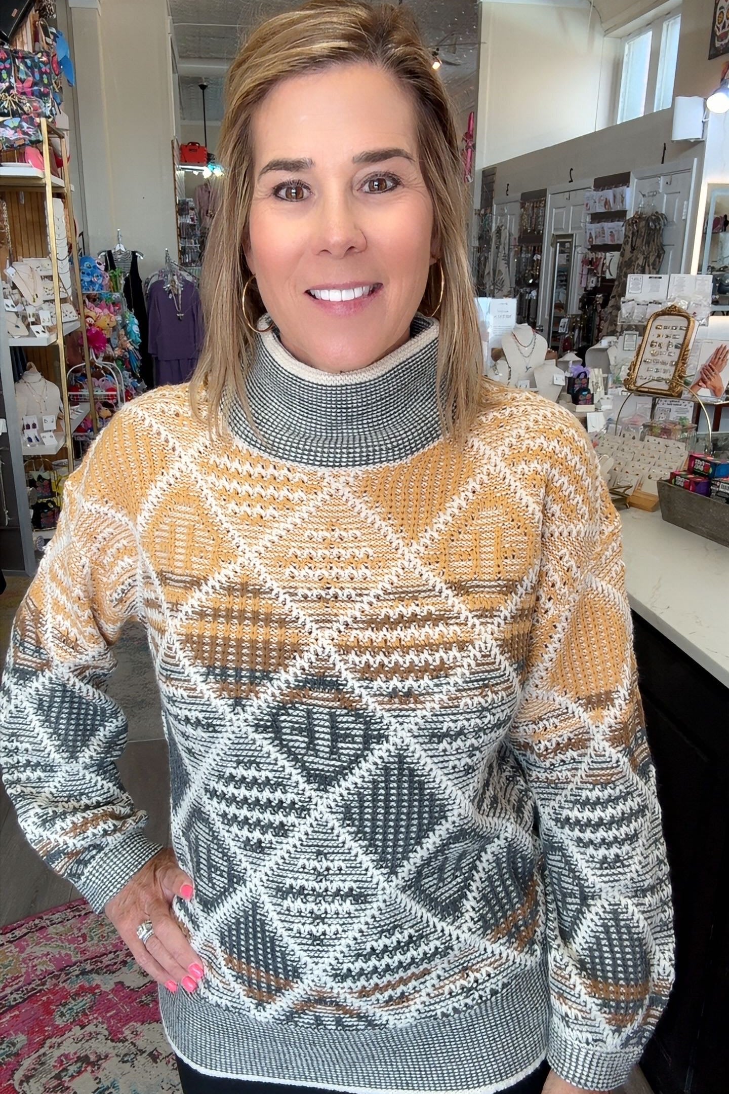 A woman wearing a patterned turtleneck sweater in a retail store setting. The sweater features a geometric diamond pattern in mustard yellow, gray, white, and brown.