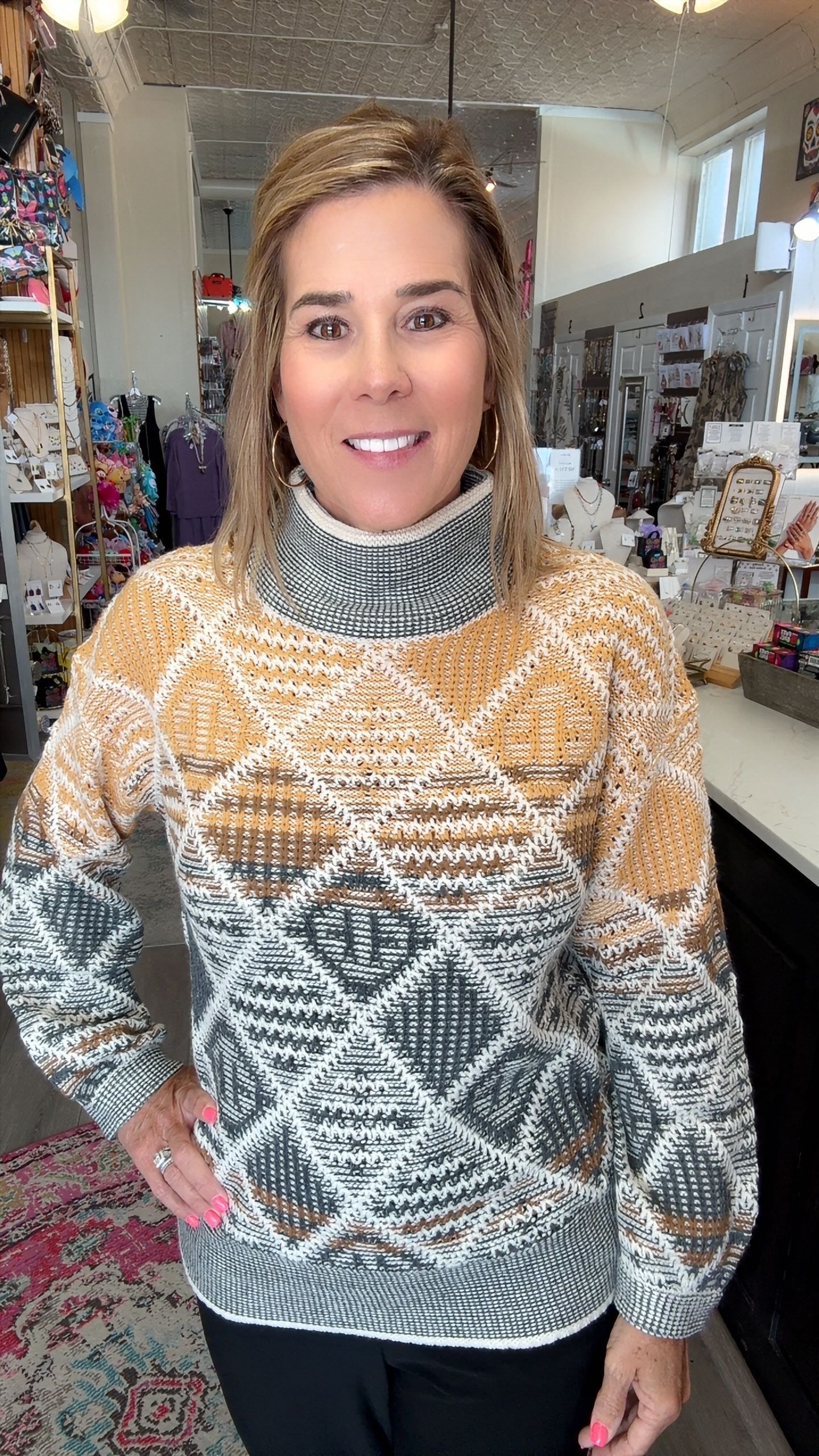 A woman wearing a patterned turtleneck sweater in a retail store setting. The sweater features a geometric diamond pattern in mustard yellow, gray, white, and brown.