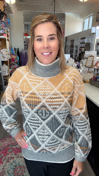 A woman wearing a patterned turtleneck sweater in a retail store setting. The sweater features a geometric diamond pattern in mustard yellow, gray, white, and brown.