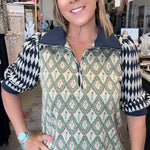 A woman modeling a patterned quarter-zip pullover top in a retail shop. The top features a cream base with green and black geometric diamond patterns on the body and sleeves, and a solid dark navy collar and zipper trim.