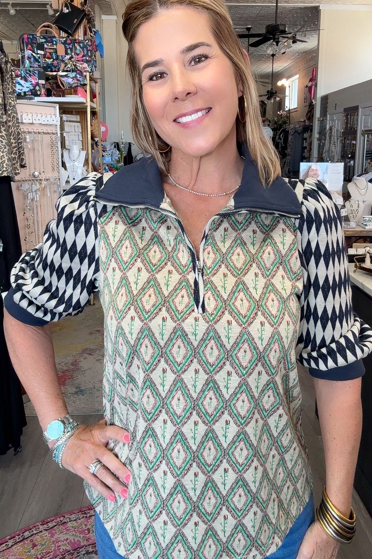 A woman modeling a patterned quarter-zip pullover top in a retail shop. The top features a cream base with green and black geometric diamond patterns on the body and sleeves, and a solid dark navy collar and zipper trim.