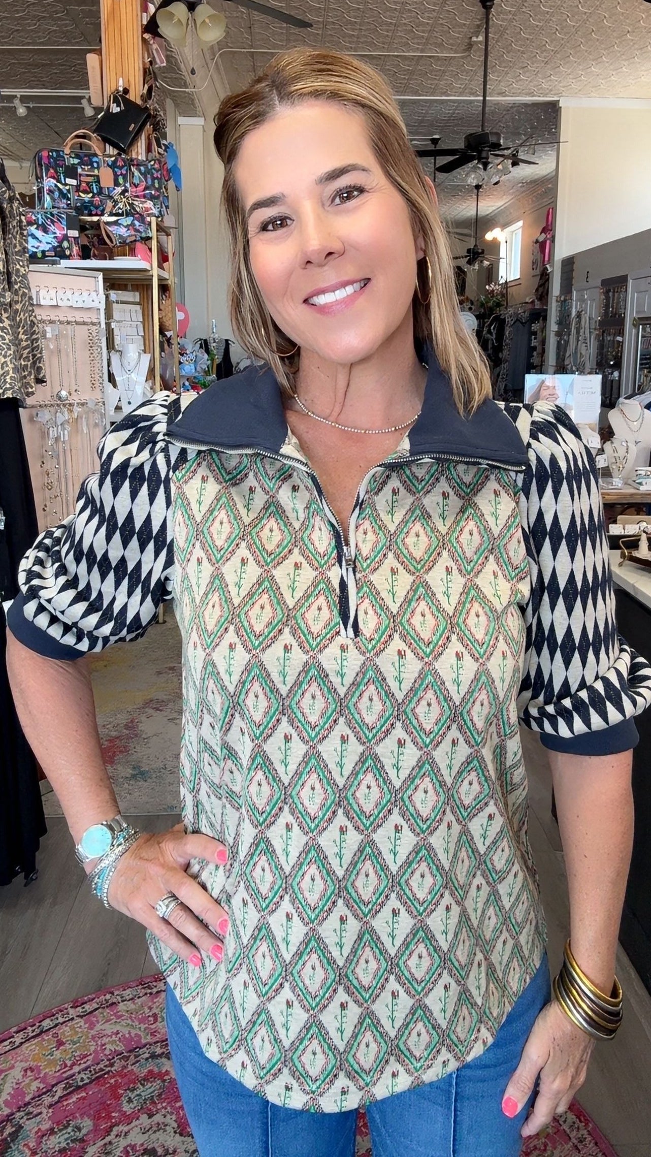 A woman modeling a patterned quarter-zip pullover top in a retail shop. The top features a cream base with green and black geometric diamond patterns on the body and sleeves, and a solid dark navy collar and zipper trim.