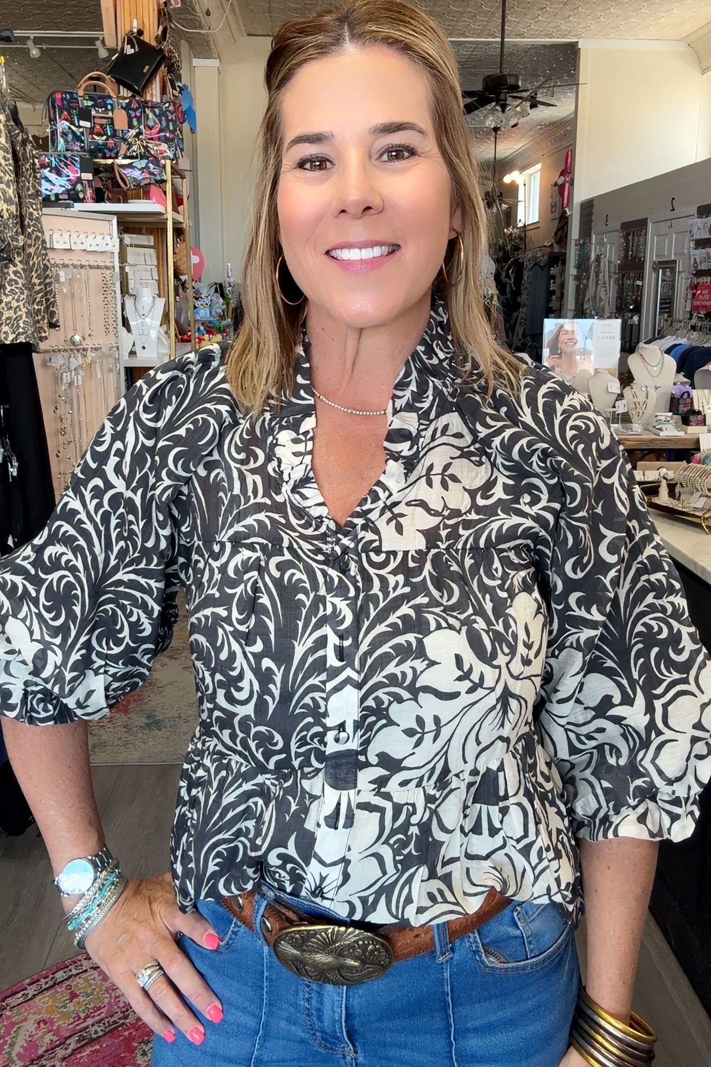 A woman wearing a black and white damask-patterned blouse with puff sleeves, styled with blue jeans and a brown leather belt with a large silver buckle, standing in a retail store.