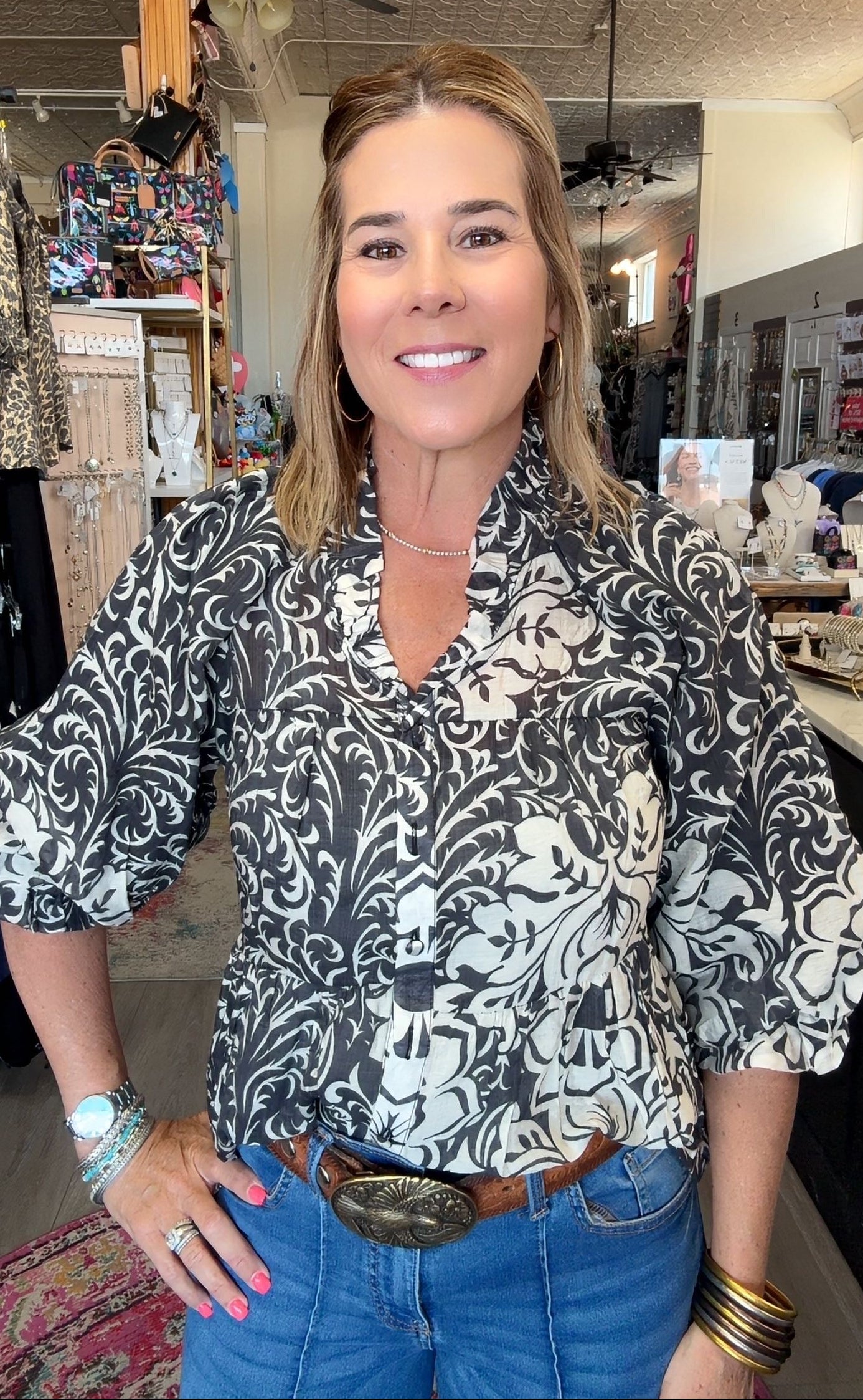 A woman wearing a black and white damask-patterned blouse with puff sleeves, styled with blue jeans and a brown leather belt with a large silver buckle, standing in a retail store.