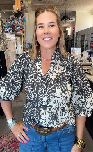A woman wearing a black and white damask-patterned blouse with puff sleeves, styled with blue jeans and a brown leather belt with a large silver buckle, standing in a retail store.