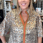 A woman modeling a leopard-print blouse with a green and orange striped collar placket, paired with blue jeans and a large silver belt buckle, standing in a retail store.